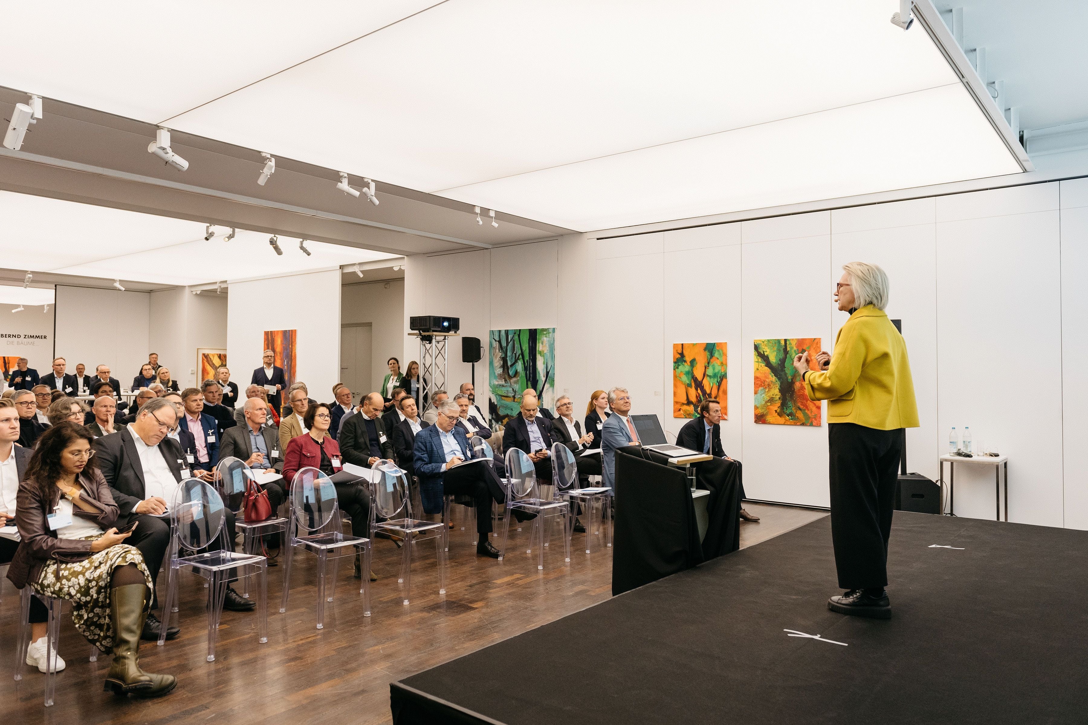 Das Bild zeigt eine Frau, die auf einer Bühne vor einem aufmerksam zuhörenden Publikum spricht. Der moderne Konferenzraum ist hell beleuchtet, mit abstrakten Gemälden an den Wänden und transparenten Stühlen für die Zuhörer.
