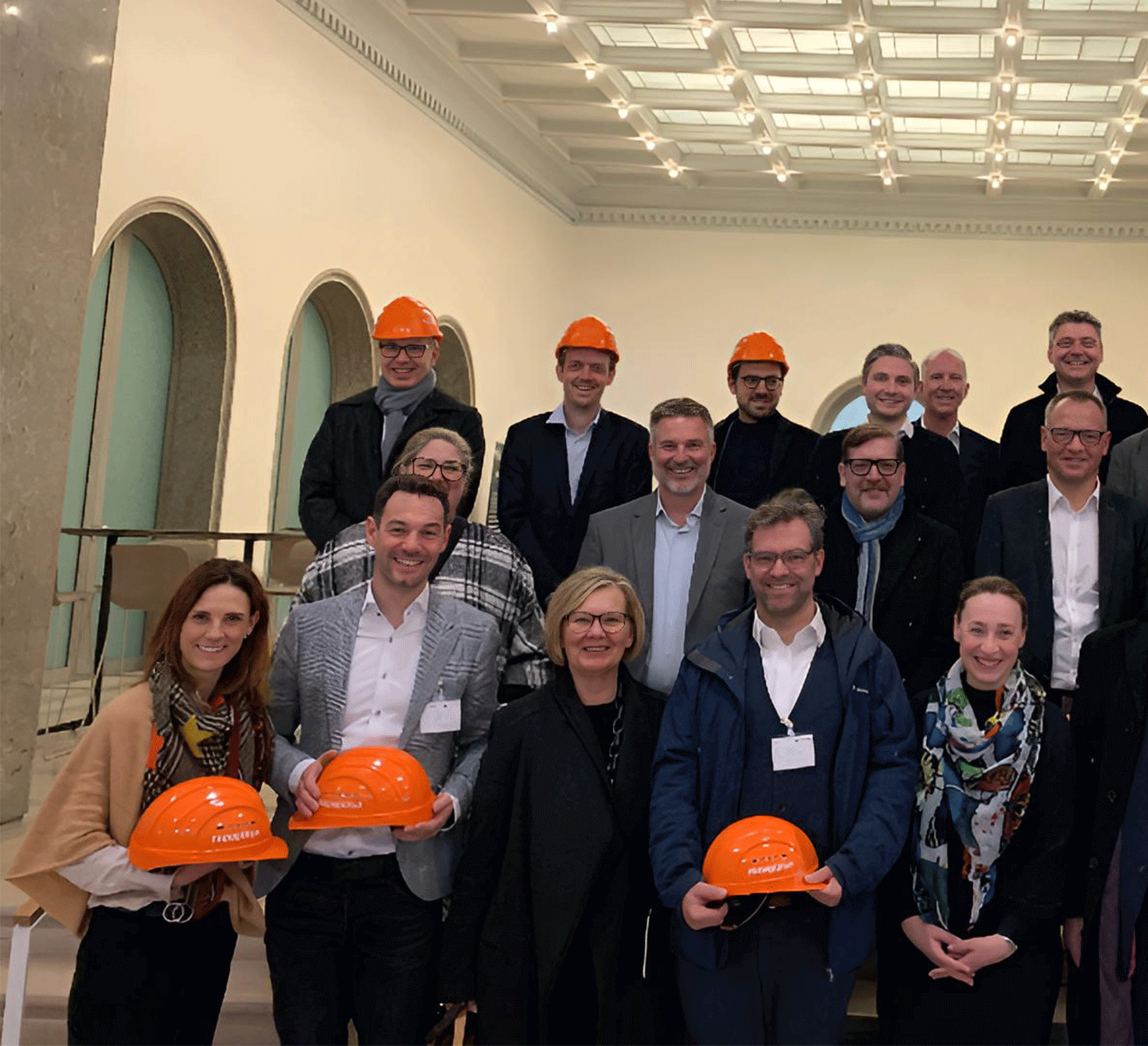 Das Bild zeigt eine Gruppe von geschäftlich gekleideten Menschen in einem Eingangsbereich. Einige von ihnen halten orangefarbene Bauhelme in der Hand.