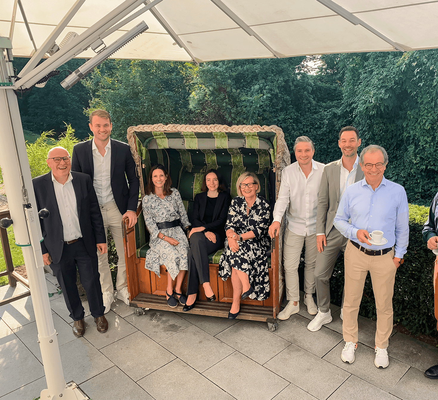 Das Bild zeigt acht Personen, die entspannt auf einer Terrasse im Garten lachen. Drei Frauen sitzen in einem grünen Strandkorb, während fünf Männer – zwei links, drei rechts – um sie herumstehen. Alle tragen formelle bis smart-casual Kleidung.