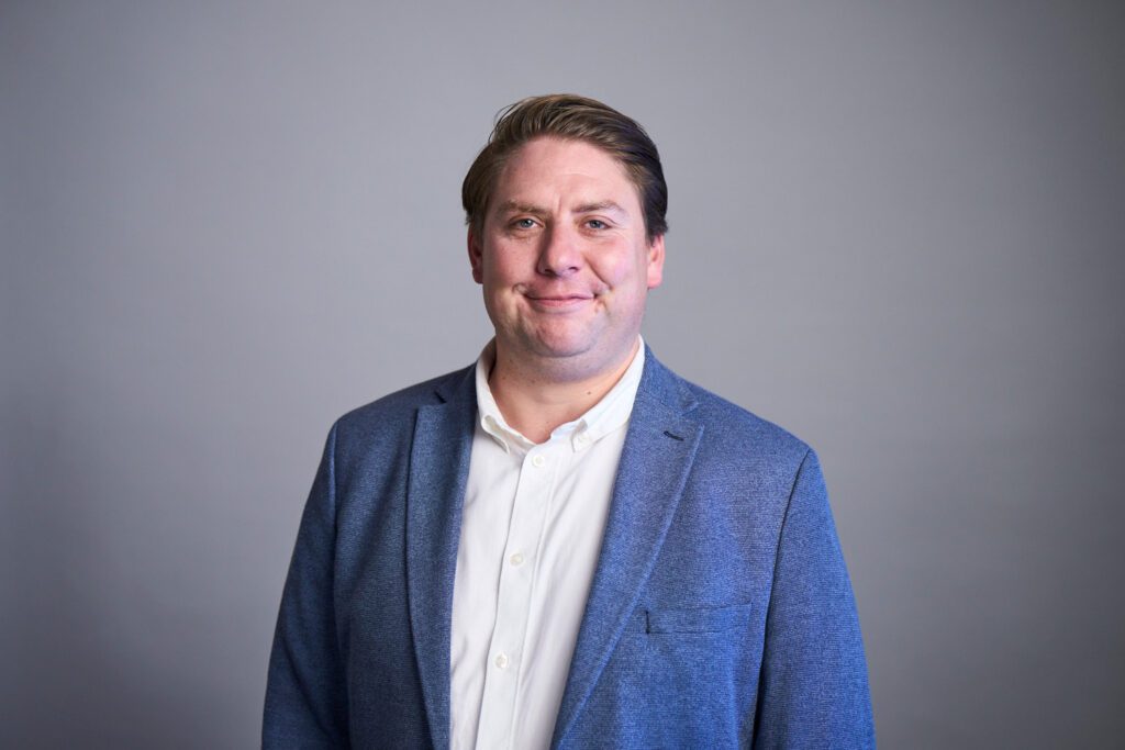 Studio portrait of a smiling H&Z consultant with swept-back hair in a blue blazer and white shirt against a grey background | H&Z
