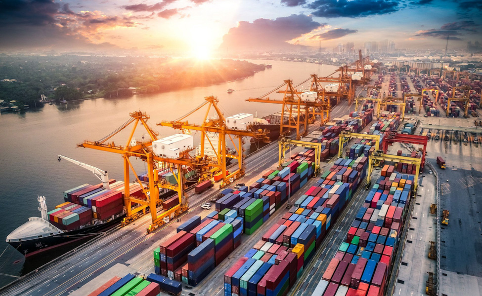 Aerial sunrise view of a busy container port with gantry cranes, ships and stacks of containers along a river | H&Z