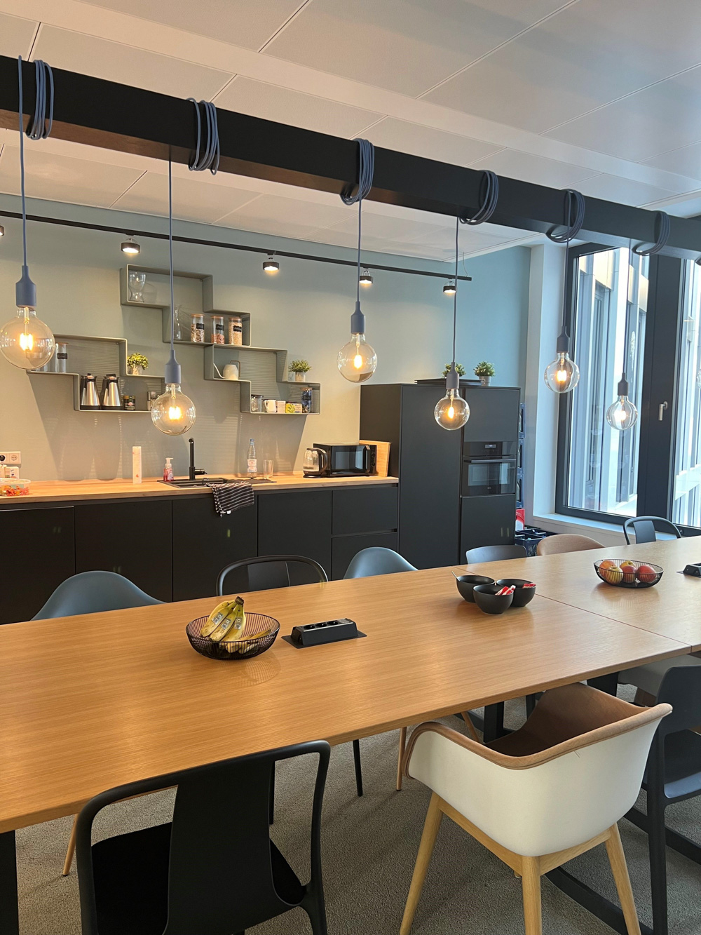 Modern office kitchen and meeting area with hanging lights, sleek cabinets, and a long wooden table set with fruit, reflecting H&Z's collaborative workspace and corporate culture.