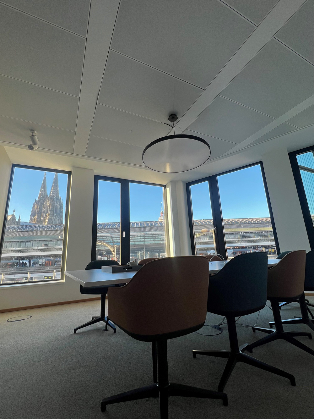 Meeting room with round table and brown chairs positioned beside panoramic windows overlooking Cologne Cathedral and central station, illustrating H&Z's inspiring workspace for client collaboration.