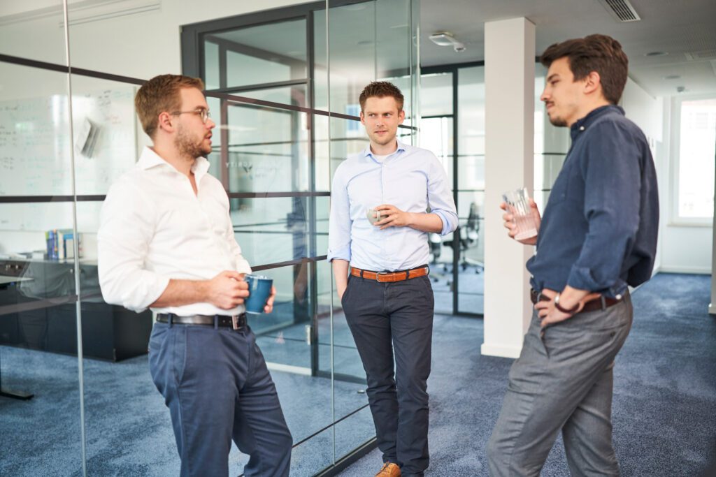 Three male consultants in a modern glass-walled office engaged in conversation while holding coffee cups, illustrating collaborative discussion and partnership at H&Z.