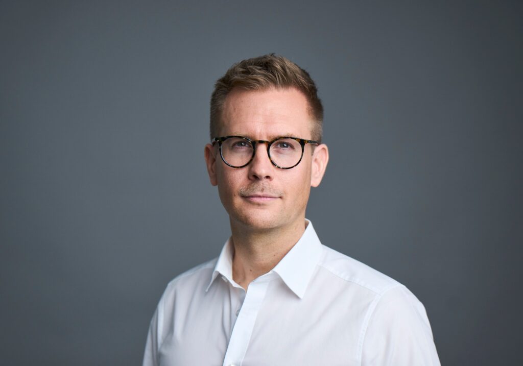 Professional male consultant wearing a white shirt and glasses, posing confidently against a neutral grey background, symbolizing expertise and professionalism.