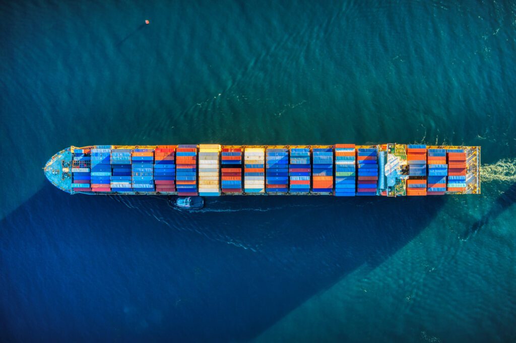 Aerial view of a container ship carrying colorful cargo boxes sailing through deep blue water – HZ Group consulting logistics and freight industry.