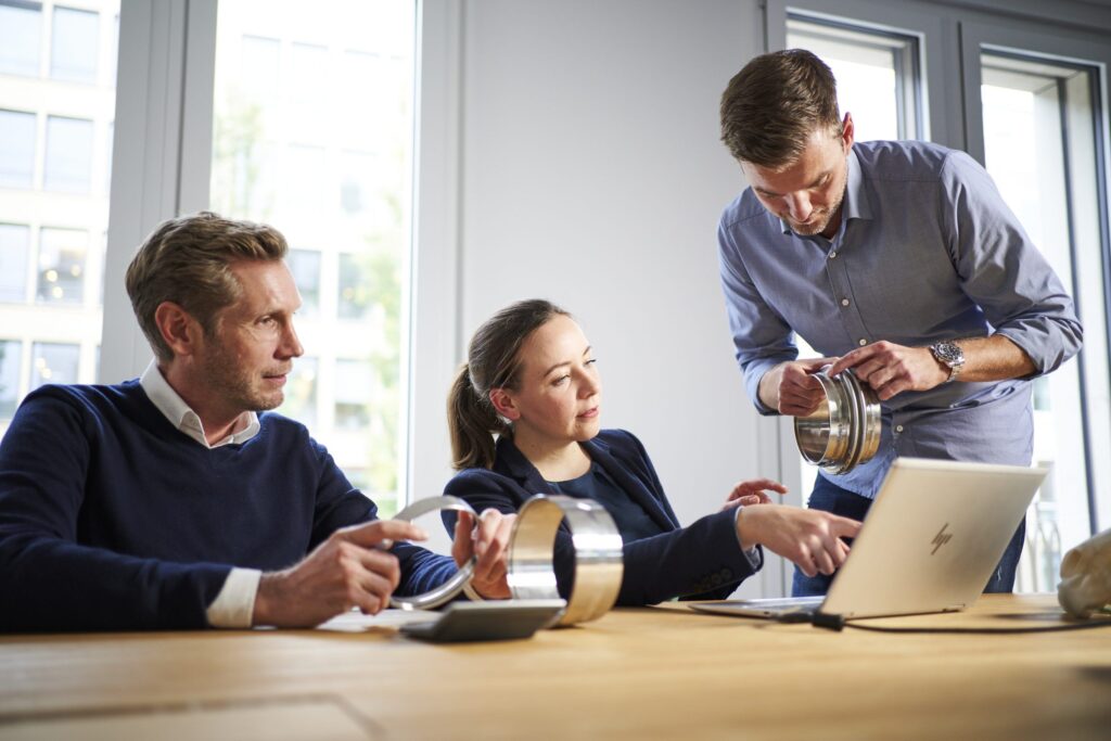 Consulting team meeting in a modern office: two men and a woman examining product samples and discussing a project around a table with a laptop.