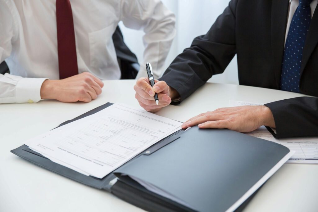 Close-up of business professionals in suits signing a contract on a clipboard, representing purchasing cooperations, procurement agreements, and collaborative consulting.