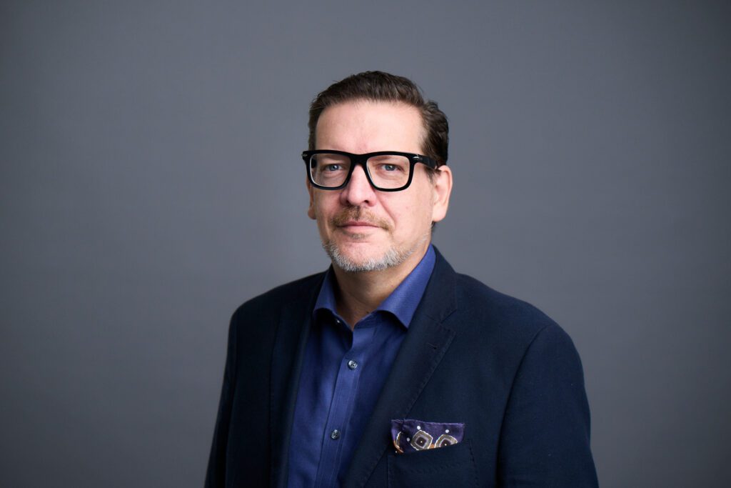 Portrait of a middle-aged man with dark-framed glasses and trimmed beard wearing a navy blue suit and pocket square, portraying confidence and strategic leadership as a consulting professional.