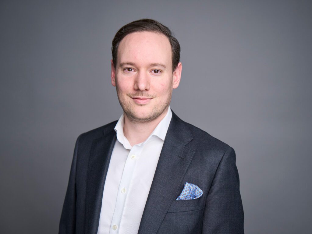 Professional headshot of Soenke Mehrtens, male consultant wearing a dark grey suit with white shirt and blue pocket square, smiling confidently against a grey background, representing strategic consulting expertise.