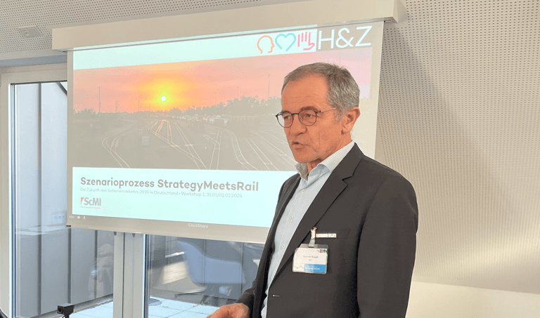 Senior male consultant in a dark suit and glasses giving a presentation during the “Strategy Meets Rail” scenario process seminar, standing beside a projector screen displaying an orange-hued railway network at sunrise, illustrating supply chain strategy and rail sector consulting.