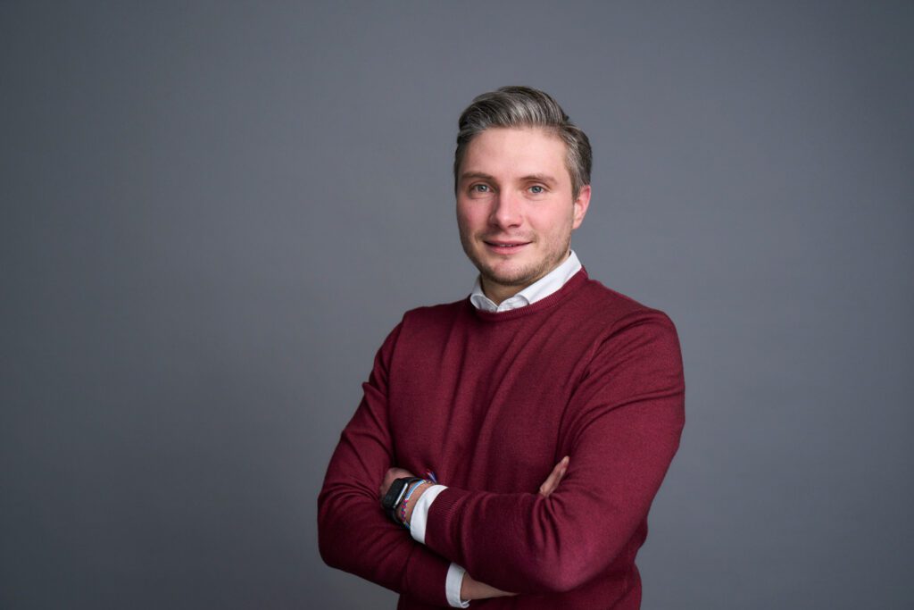 Professional portrait of Sven Steinert, a young consultant wearing a maroon sweater and white shirt with arms crossed, representing dynamic leadership in procurement consulting.
