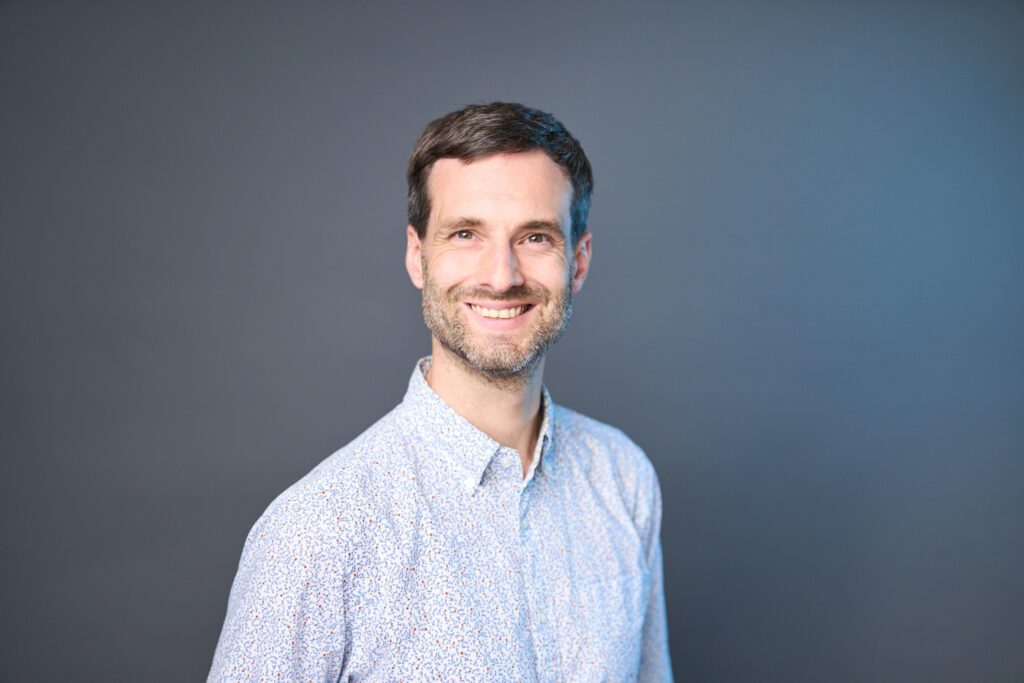 Studio portrait of an H&Z consultant with short dark hair and beard in a patterned light blue shirt | H&Z