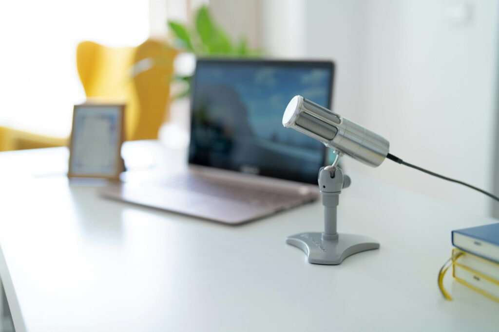 Desk setup featuring a sleek microphone on a stand with a blurred laptop, tablet, and plant in the background, representing remote consulting, webinars, and professional communication in a modern office