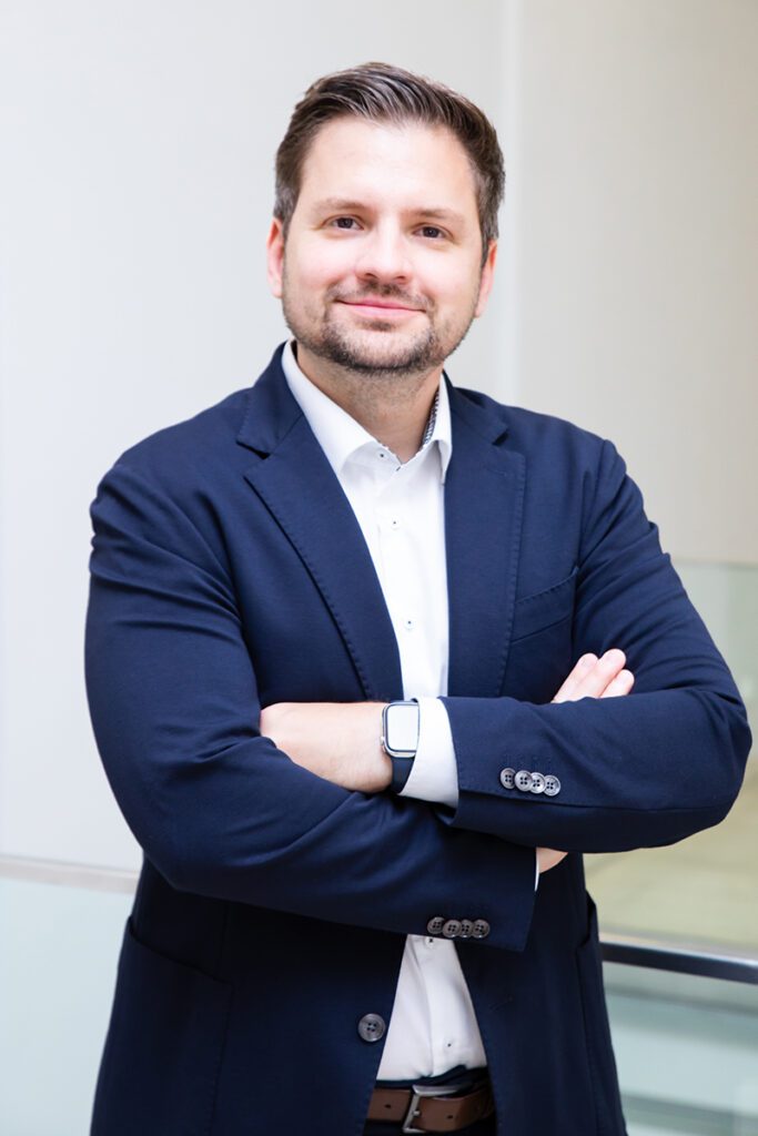 Portrait of Michael Schuh, professional consultant at H&Z, smiling with arms crossed in a navy blue suit, representing leadership and expertise.