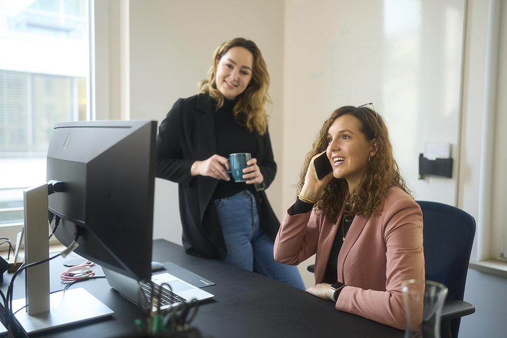 H&Z consulting colleagues collaborating in a modern office; one woman speaks on the phone at a desk while another stands with a coffee cup, illustrating teamwork and client communication.