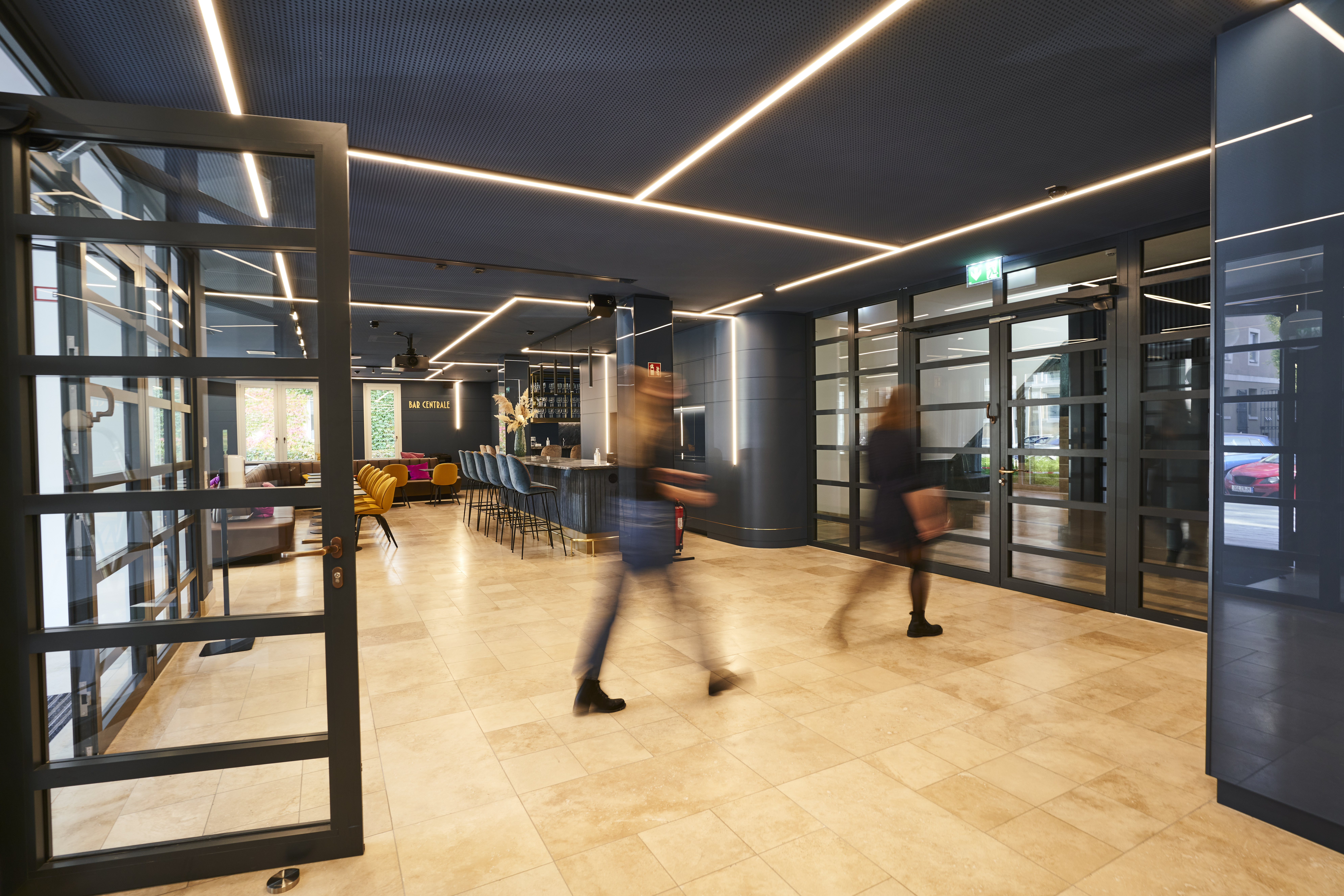 Blurred employees walking through a modern office lounge with bar area, beige tile floors and large glass doors, representing dynamic consulting workspace and hospitality environment