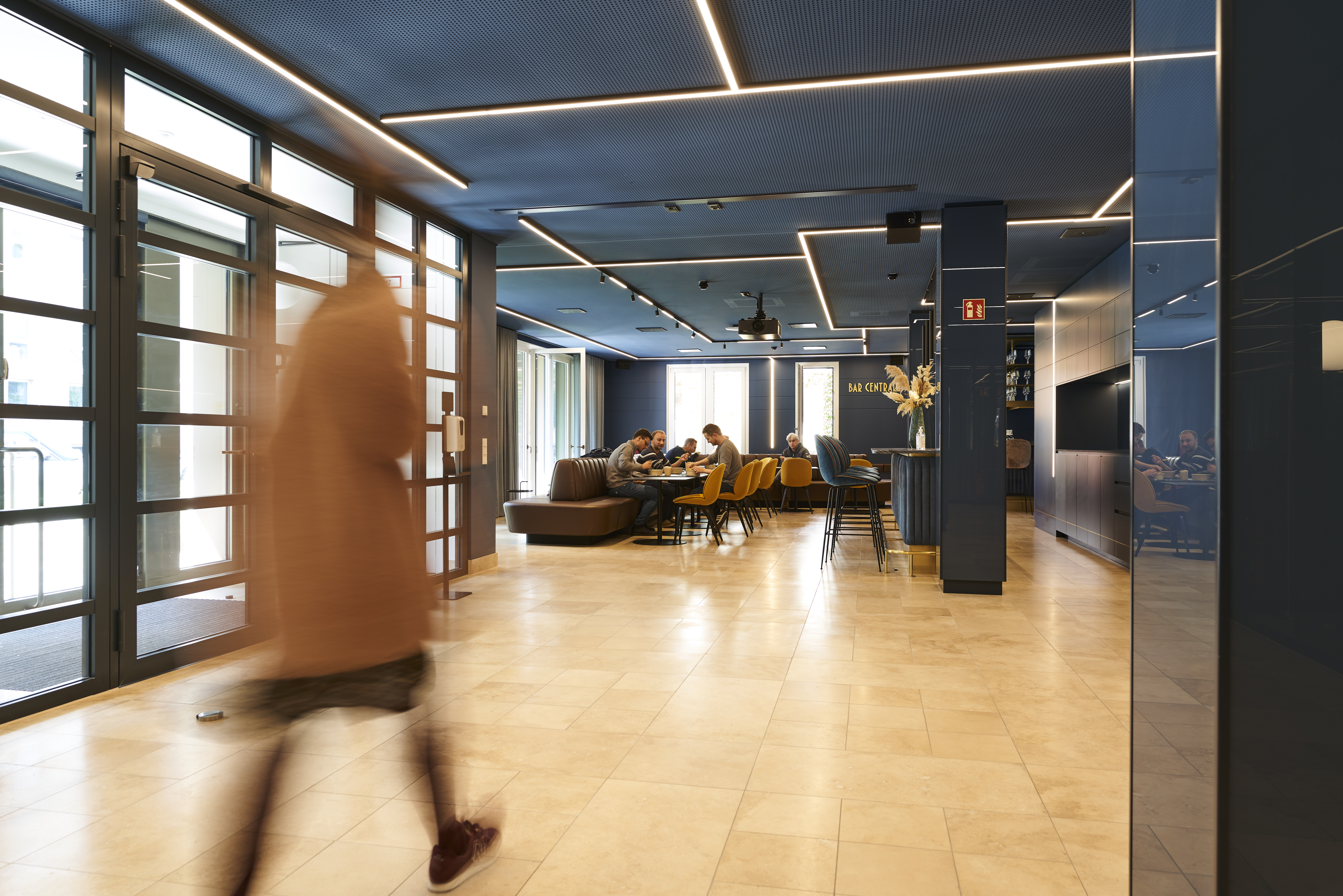 Modern consulting office lobby with a blurred figure walking across polished floor and a meeting area with colleagues in the background, conveying dynamic business environment and collaboration.