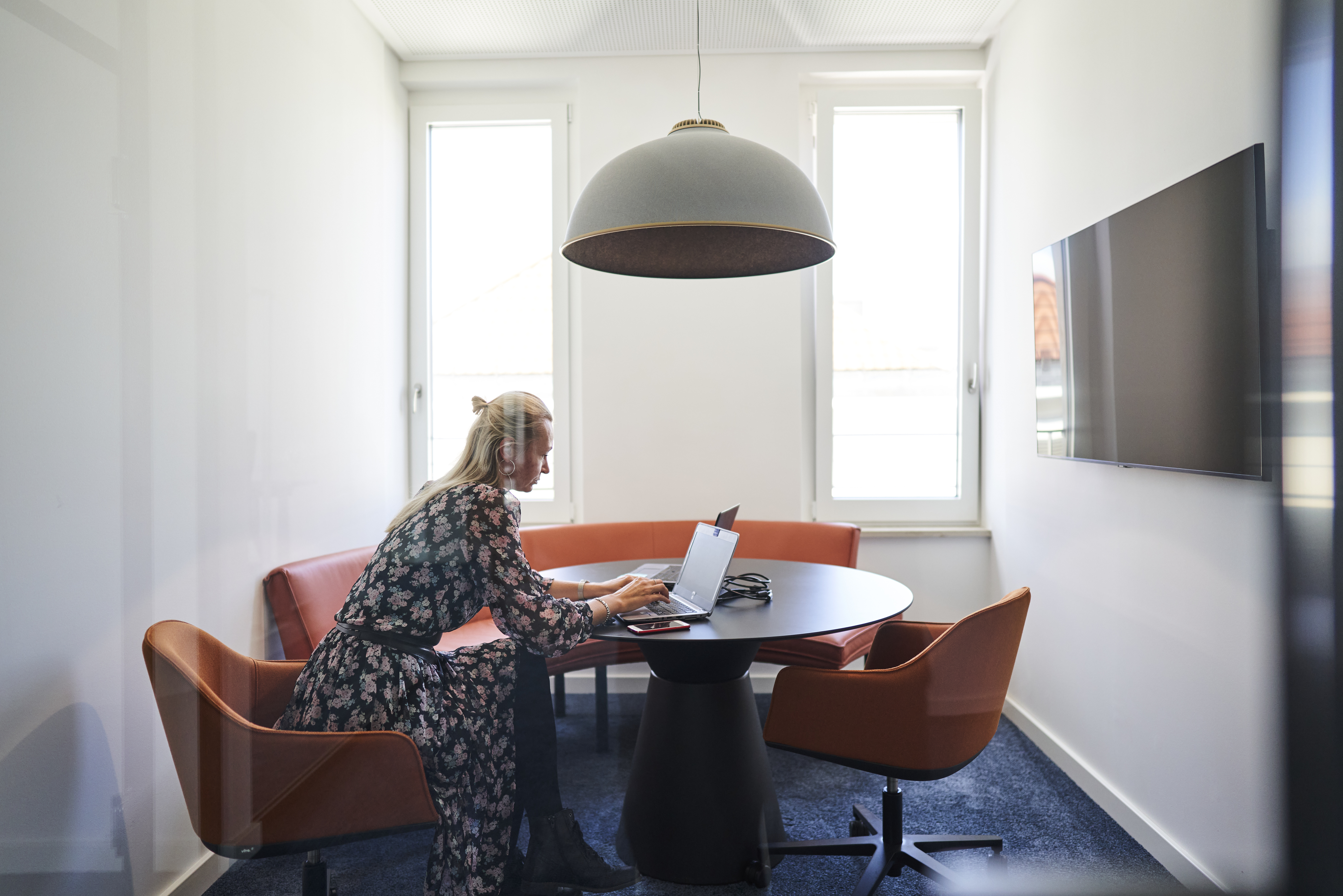 Female consultant working on laptop at small meeting table with monitor in modern office meeting room