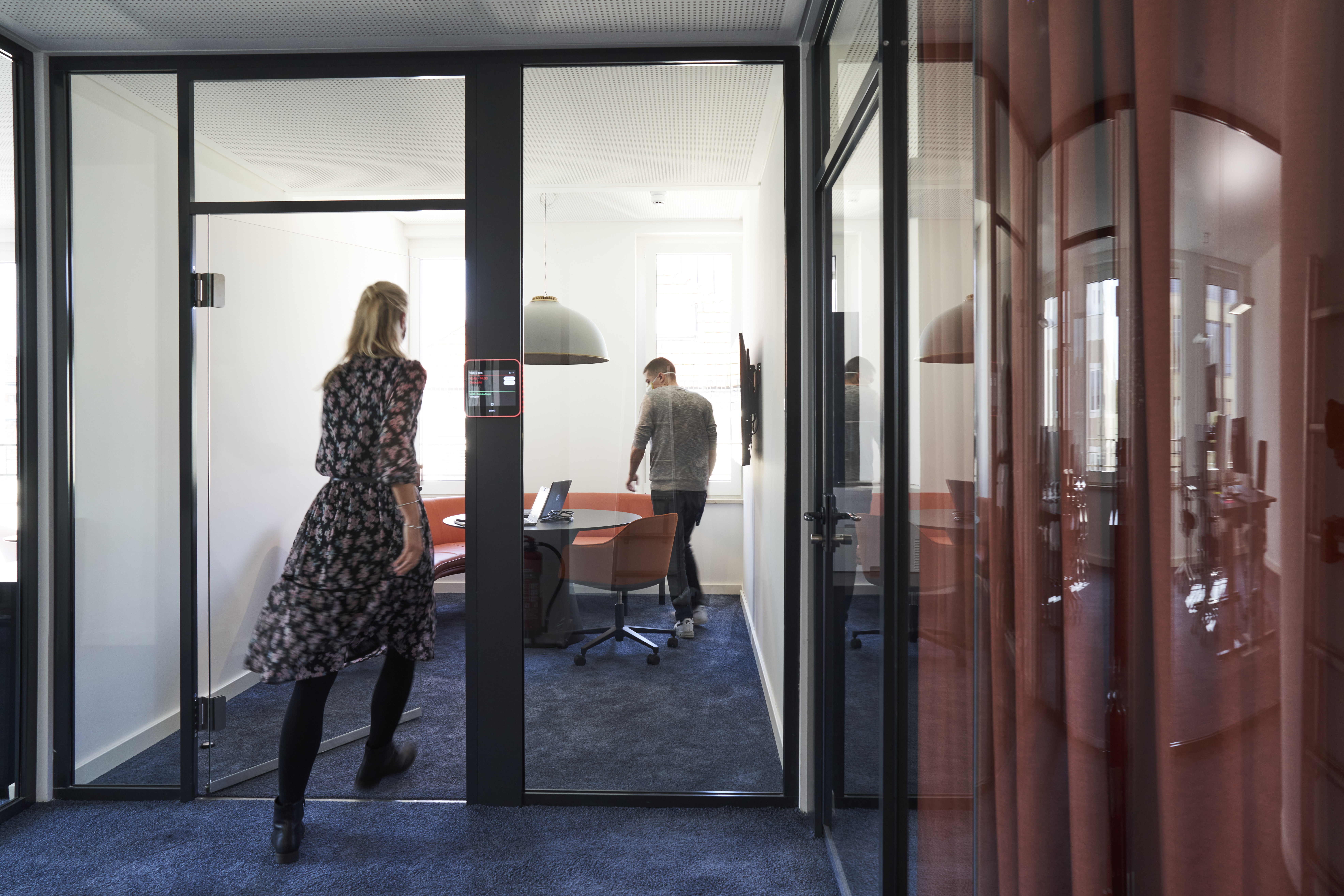 Consultant in patterned dress walking through glass meeting room while colleague works at round table in modern office, illustrating dynamic consulting collaboration