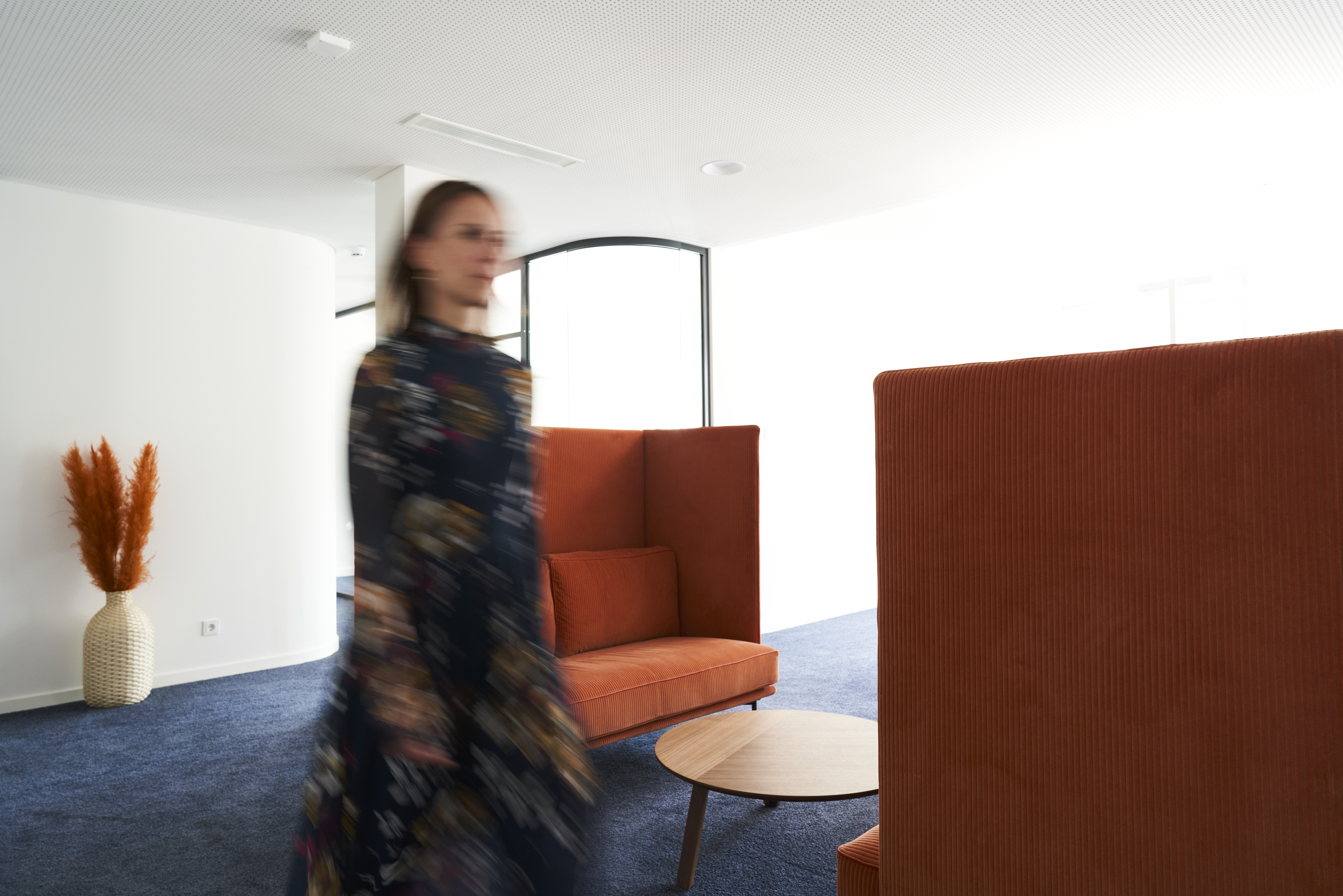 Blurred female consultant walking through modern office space with high-back lounge chairs and coffee table representing dynamic work environment
