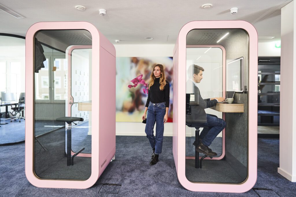 Two pink acoustic phone booths in a modern office; a male consultant works on his laptop inside while a female colleague stands outside, representing focus and collaboration spaces in a consulting firm