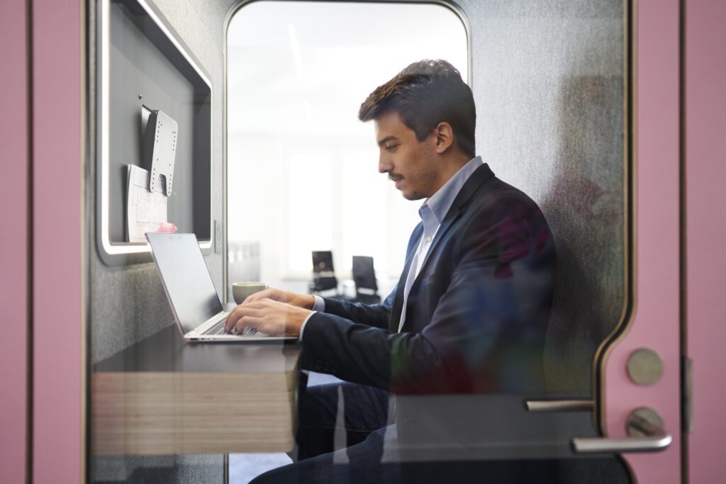 Male consultant working on laptop inside pink acoustic phone booth in modern consulting office, focusing on client project in private workspace