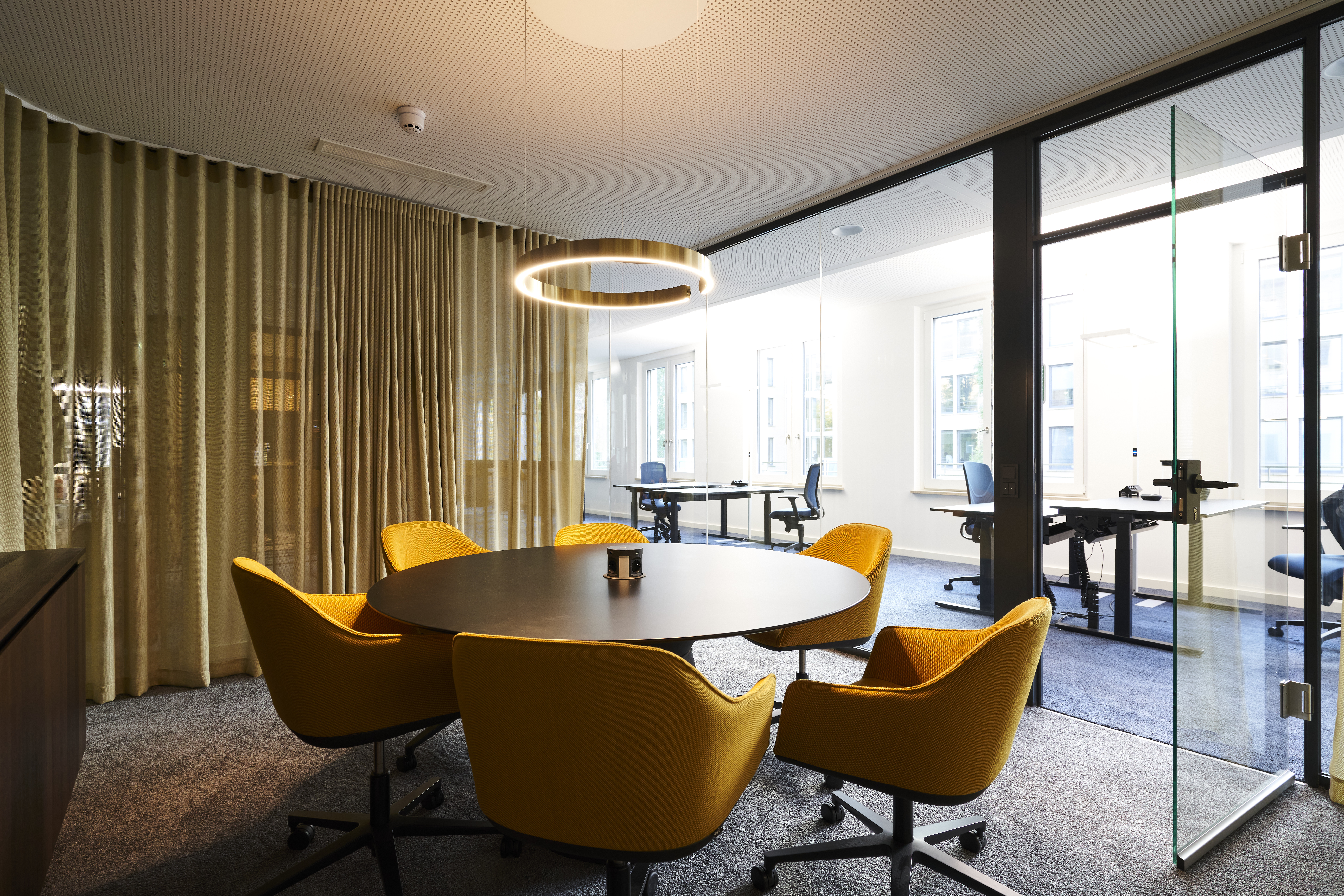 Modern meeting room with round table and yellow chairs separated by glass partition, featuring ring light and open office beyond, representing collaborative consulting workspace