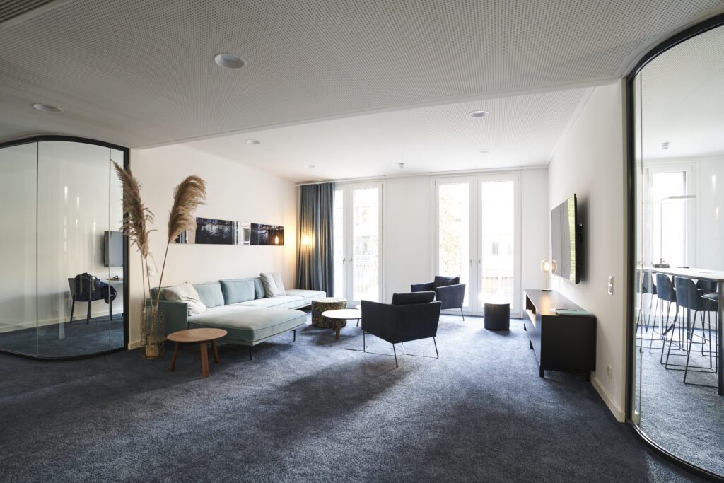 Large modern lounge area in consulting office with blue carpet, glass wall, sofa and armchairs, bar counter and large windows for client receptions and collaborative gatherings