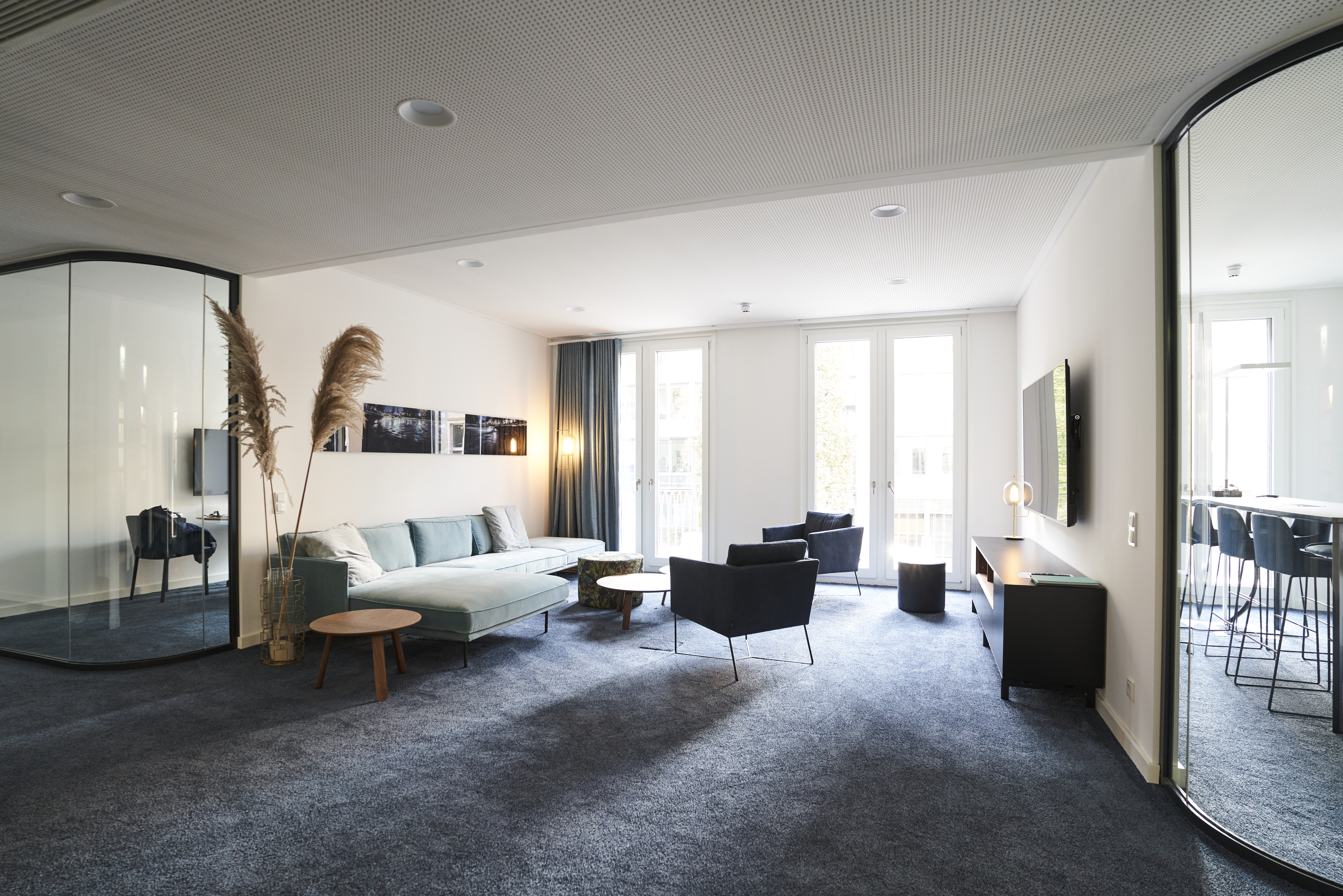 Large modern lounge area in consulting office with blue carpet, glass wall, sofa and armchairs, bar counter and large windows for client receptions and collaborative gatherings