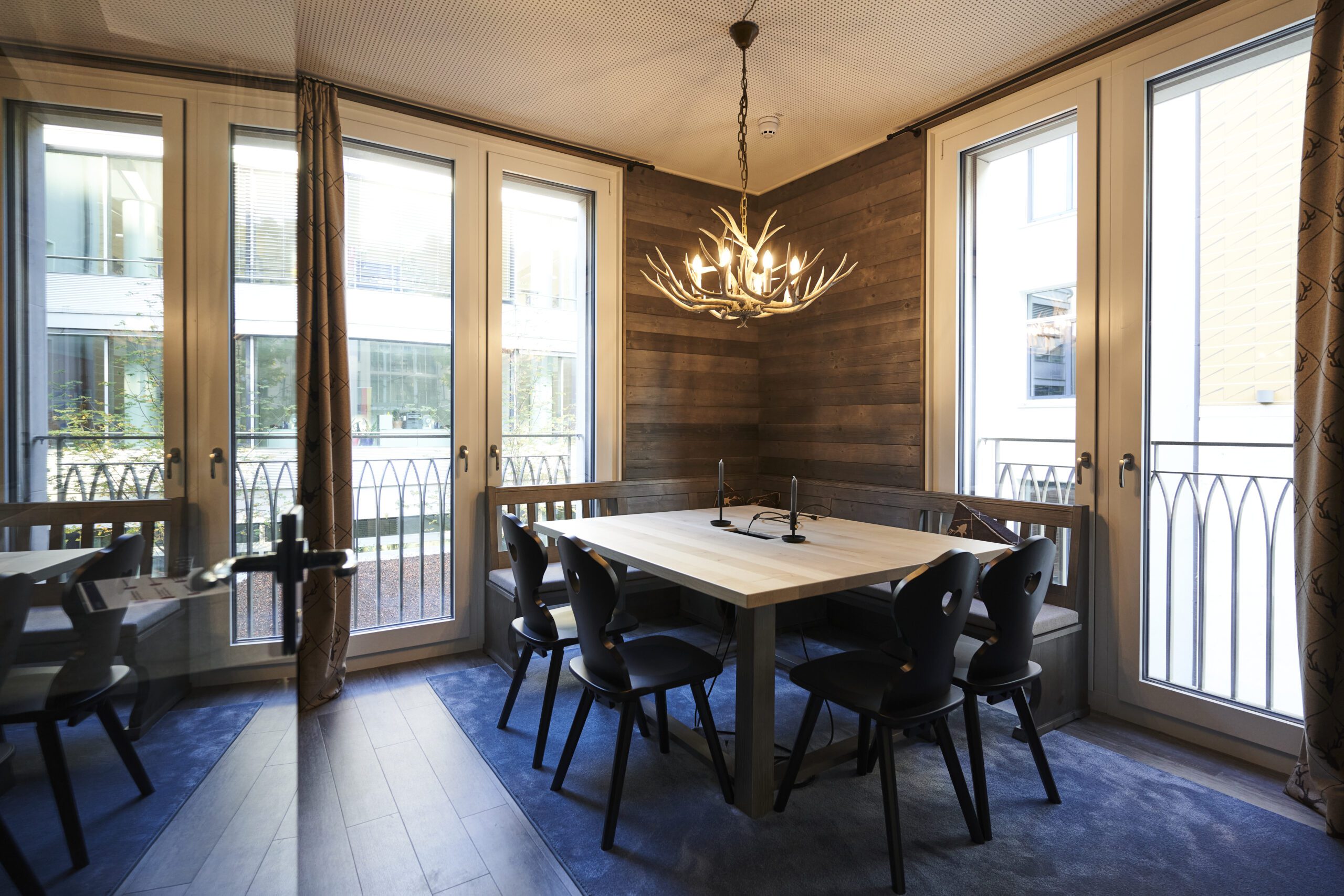 Dining or meeting area with rustic wooden table, black chairs and antler-like chandelier, set in a bright room with large windows and balcony, representing creative collaboration space in a consulting office
