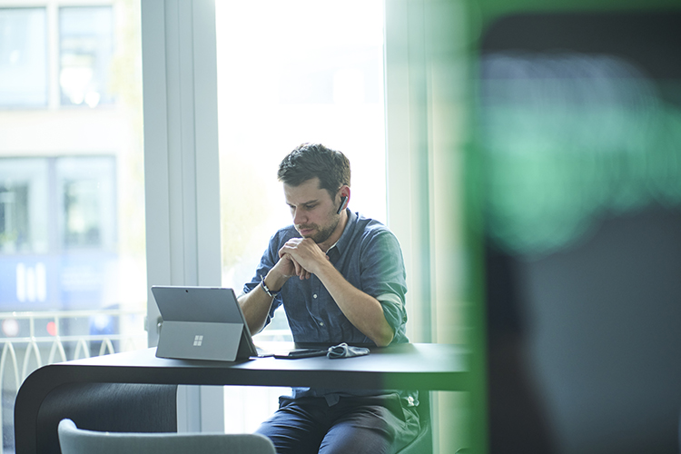 Male consultant working at a desk with headphones, using a Microsoft Surface laptop in a modern office environment, representing focused business consulting work.