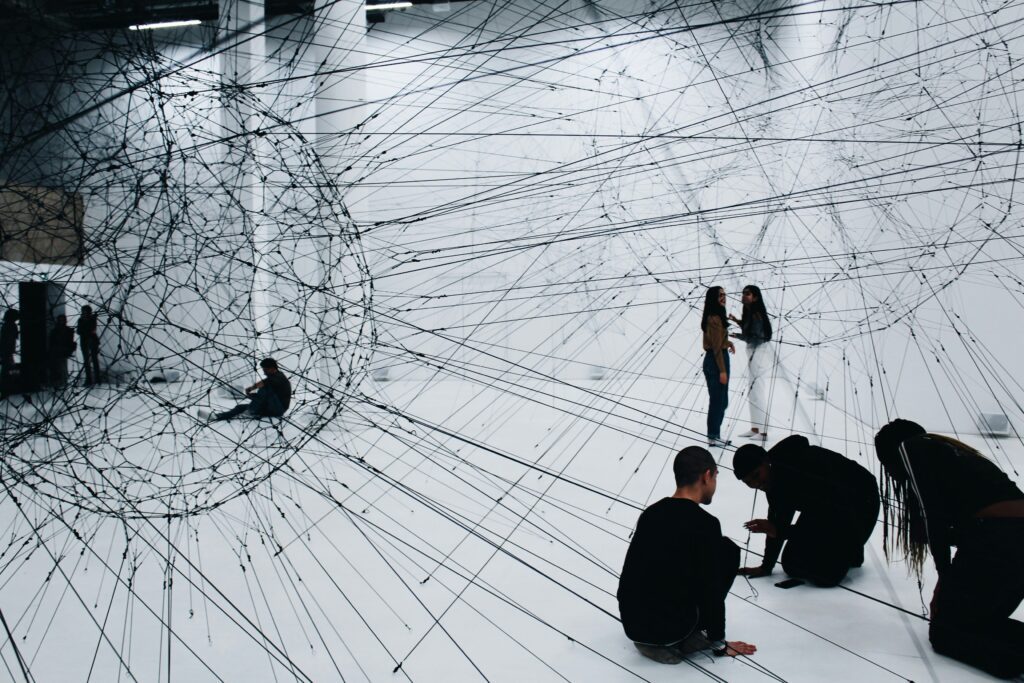 Modern art installation with people interacting among suspended strings in a spacious gallery, symbolizing complex networks and innovative thinking in consulting projects.