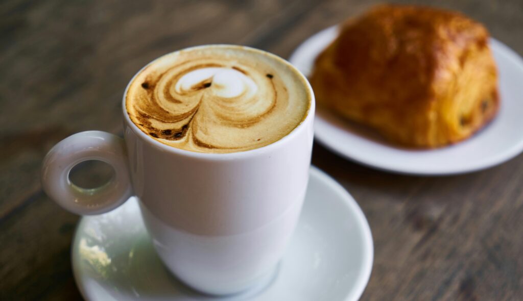 Cappuccino with latte art next to a pain au chocolat pastry on a wooden café table | H&Z