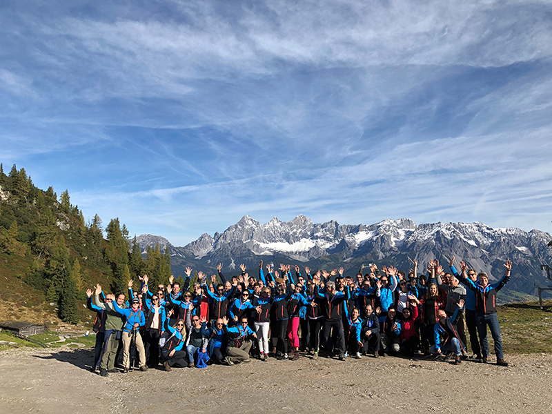 H&Z consulting team group photo raising hands during an outdoor retreat in the mountains, symbolizing teamwork and team spirit against an alpine landscape.