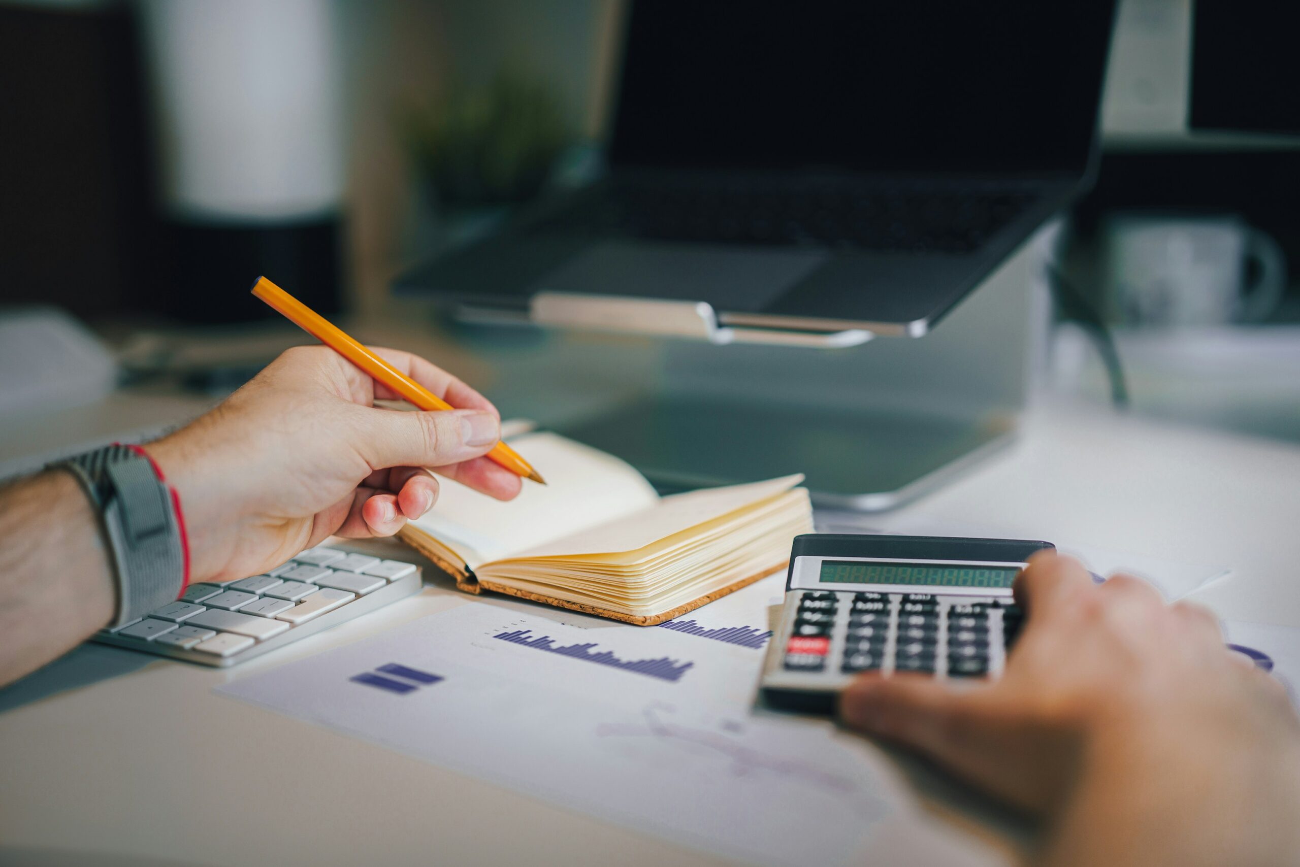 Hand of a consultant writing in a notebook with a pencil while using a calculator on top of financial documents, illustrating budgeting and cost analysis services.