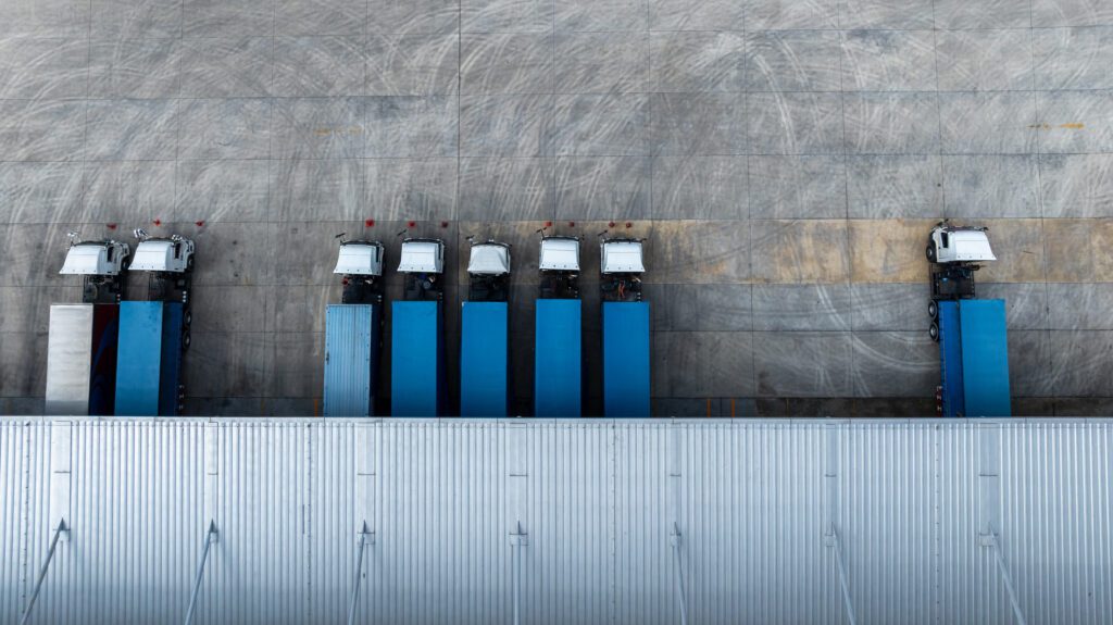 Aerial view of blue semi trucks lined up at a warehouse loading dock, representing logistics efficiency and supply chain management in consulting solutions.