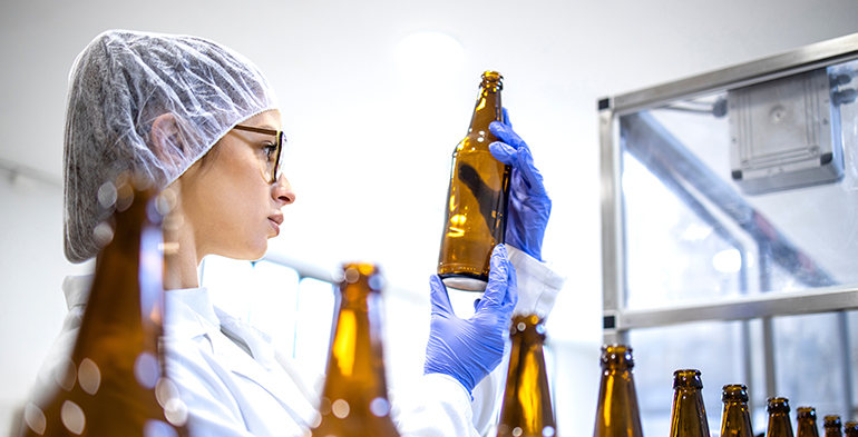 Female lab technician inspecting glass bottles in a beverage production facility, highlighting quality control and operational excellence consulting in the food and drink industry.