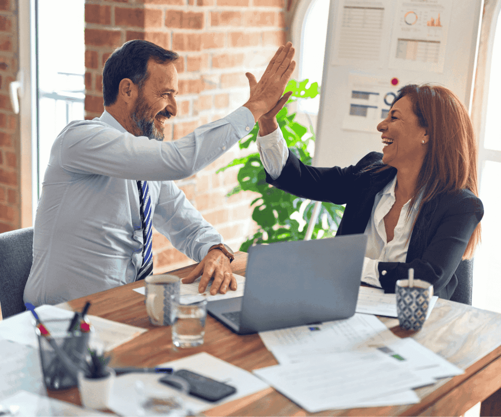 Two business professionals sharing a high-five over a workspace with laptop and documents, celebrating successful collaboration and strategic results in a consulting environment.