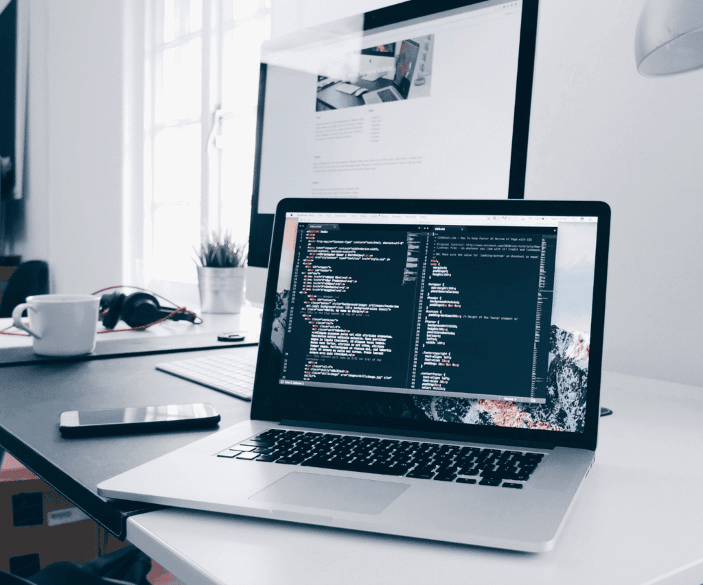 Laptop on a desk displaying lines of code with another screen in the background and office items, symbolizing software development and digital consulting workspace.