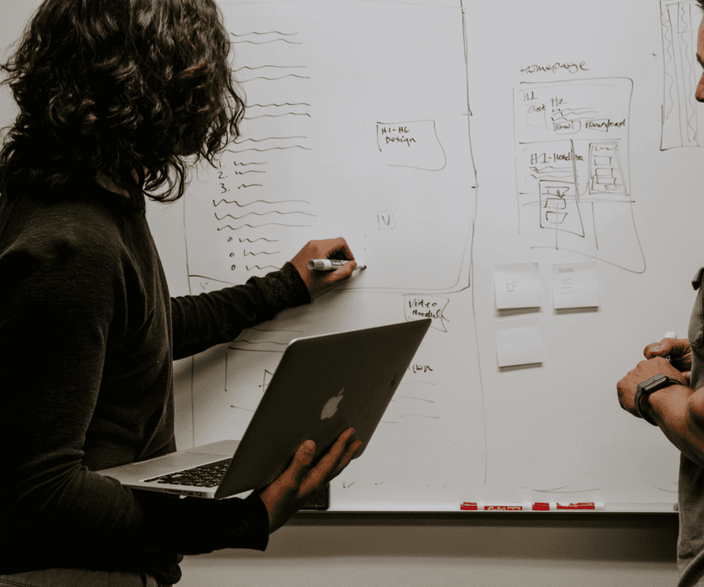 Business professionals brainstorming at a whiteboard with sketches while referencing a laptop, illustrating creative problem solving and strategic planning in consulting.