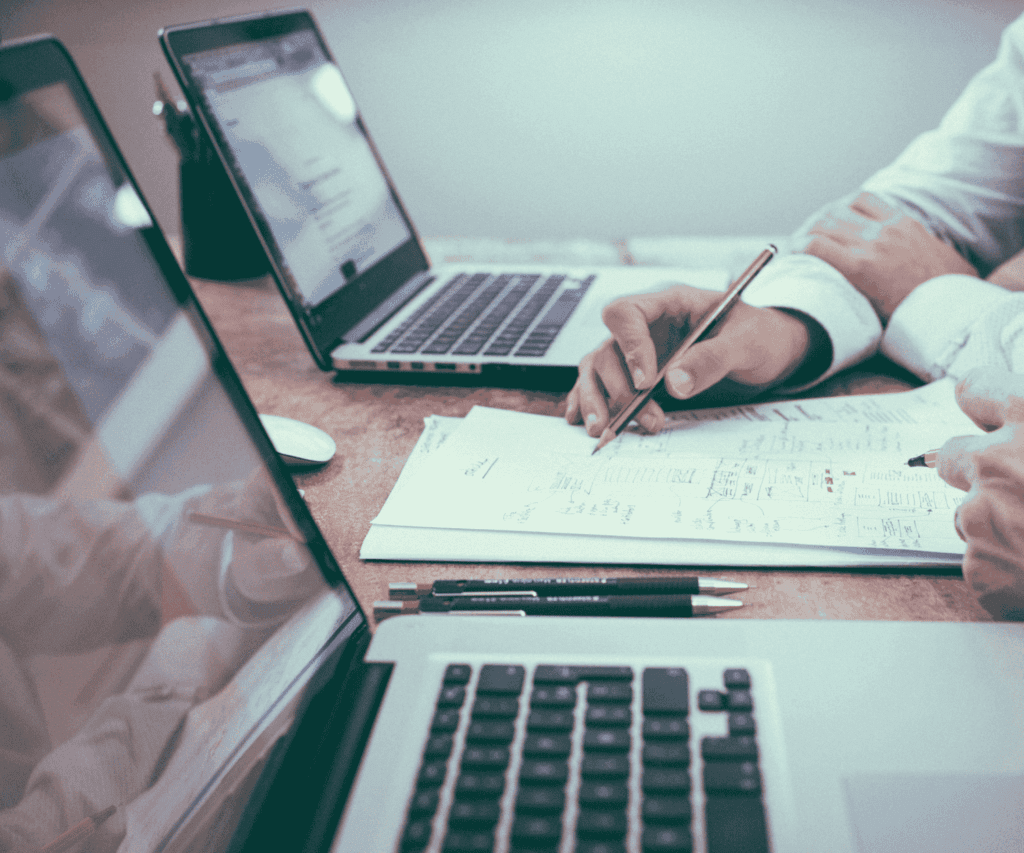 Close-up of hands taking notes on a notepad surrounded by laptops and documents, illustrating brainstorming, strategic planning, and research in consulting work.