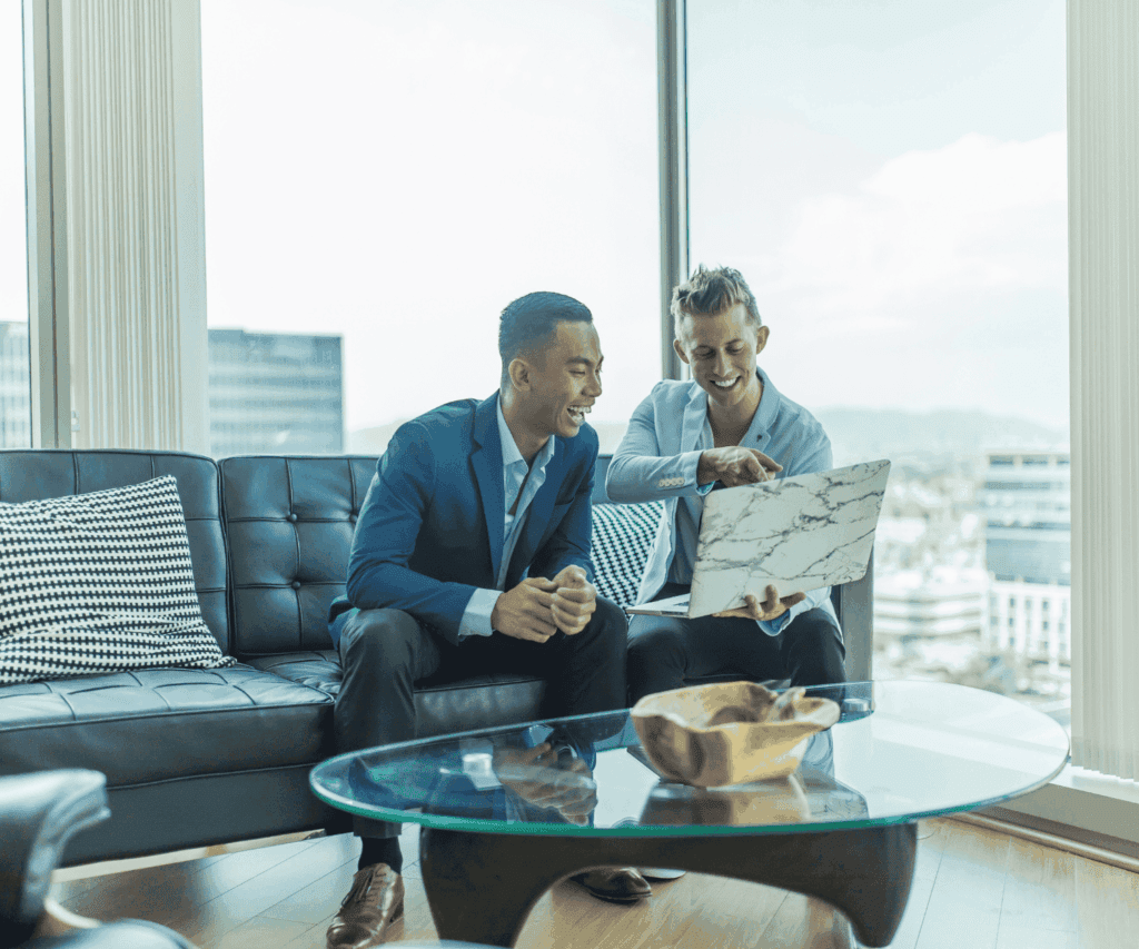 Two male professionals collaborating in a modern office lounge, reviewing a design on a laptop with a city view, symbolizing strategic consulting and teamwork.