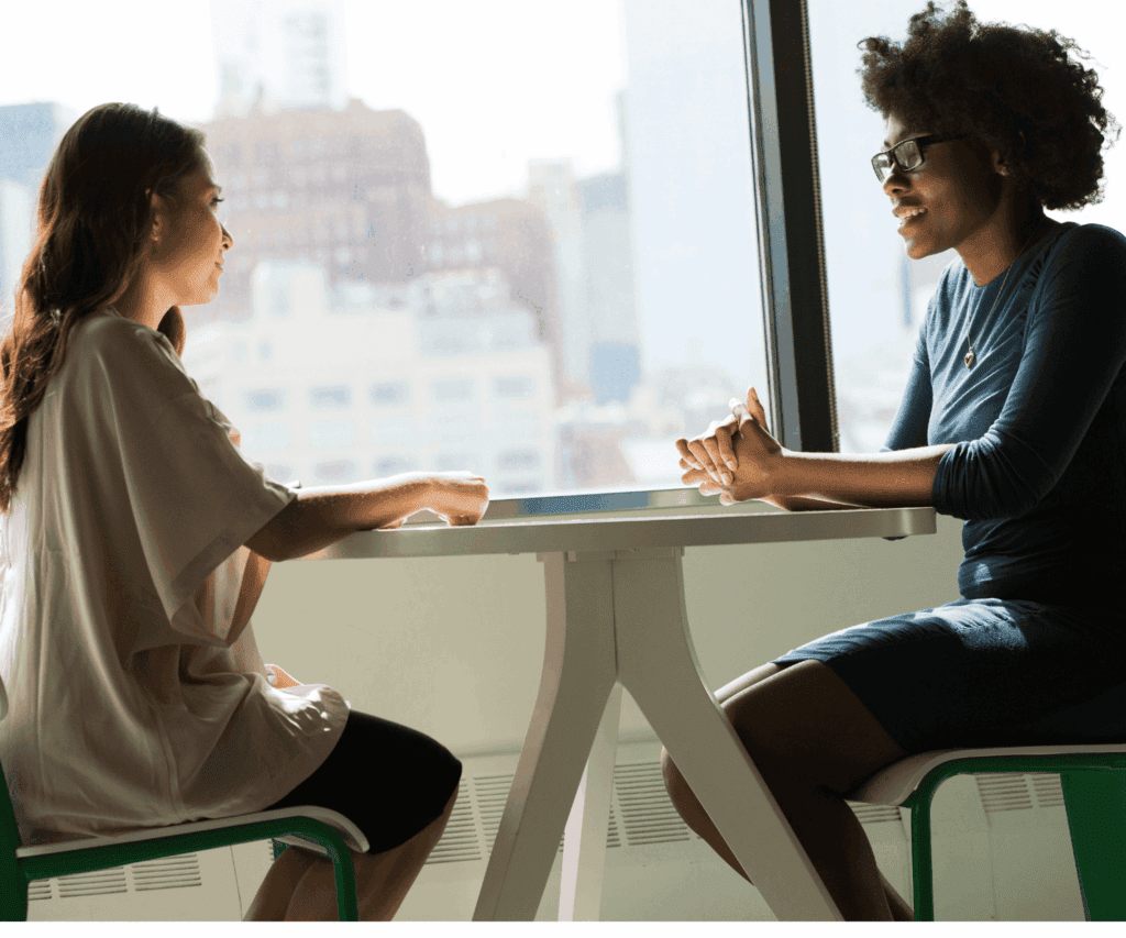 Two professional women engaged in a discussion at a table with city skyline in the background, illustrating collaborative consulting conversations and client engagement.