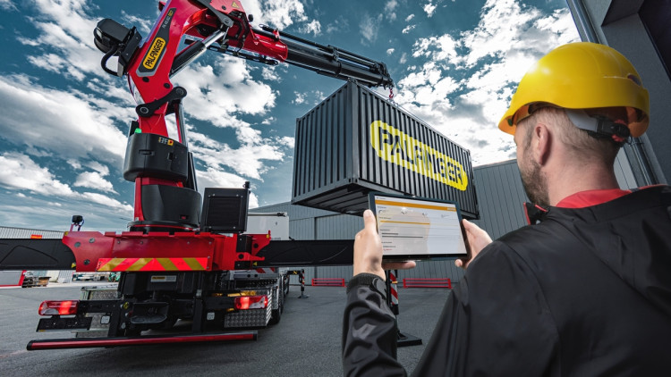 Worker using a tablet to coordinate a red crane lifting an industrial container at a shipping yard, reflecting technology-driven project management and logistics consulting.