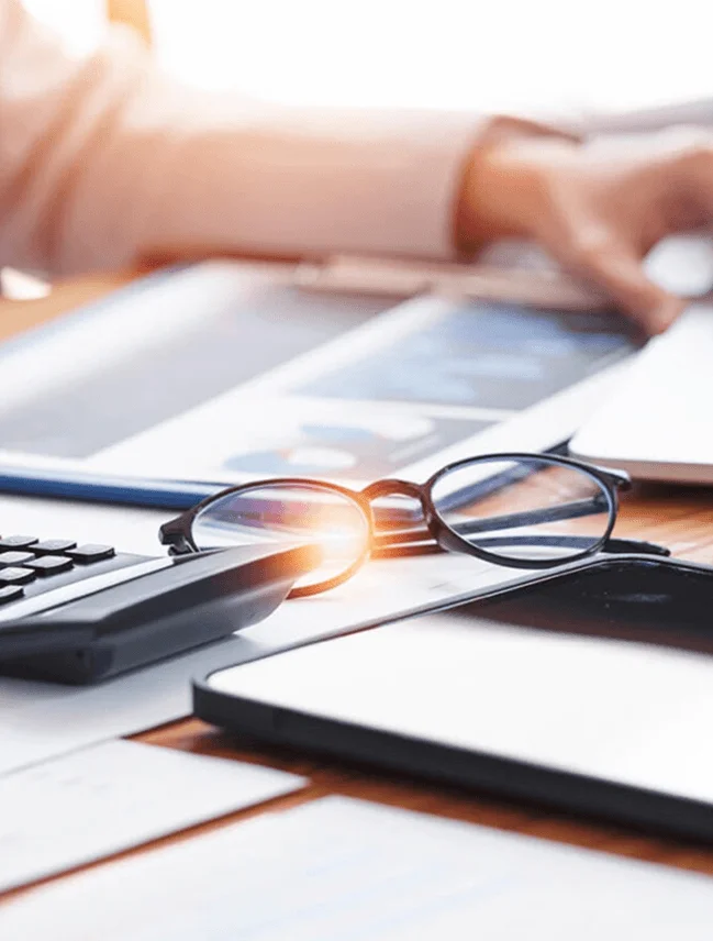 Close-up of a business workspace with eyeglasses, calculator, smartphone, and tablet on a desk with financial documents, symbolizing analysis and consulting work.