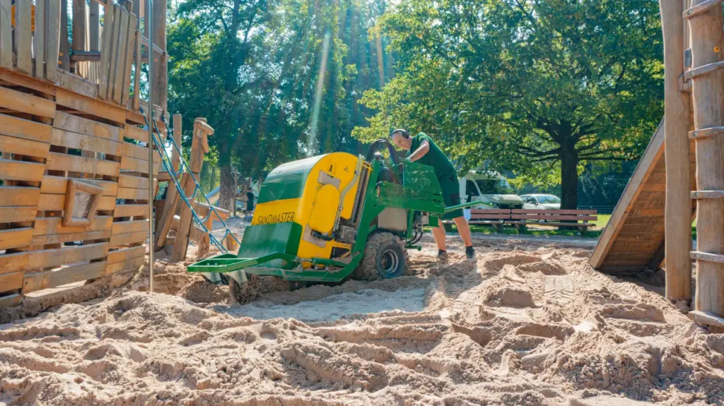 Worker operating a green and yellow construction machine in a sandy outdoor area with wooden structures and trees, representing hands-on experience and project execution in a consulting context.
