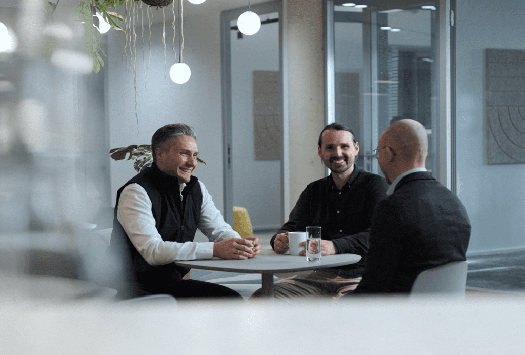 Three businessmen meeting at a round table in a modern office, smiling and discussing work over coffee, symbolizing collaboration, leadership, and consulting teamwork.
