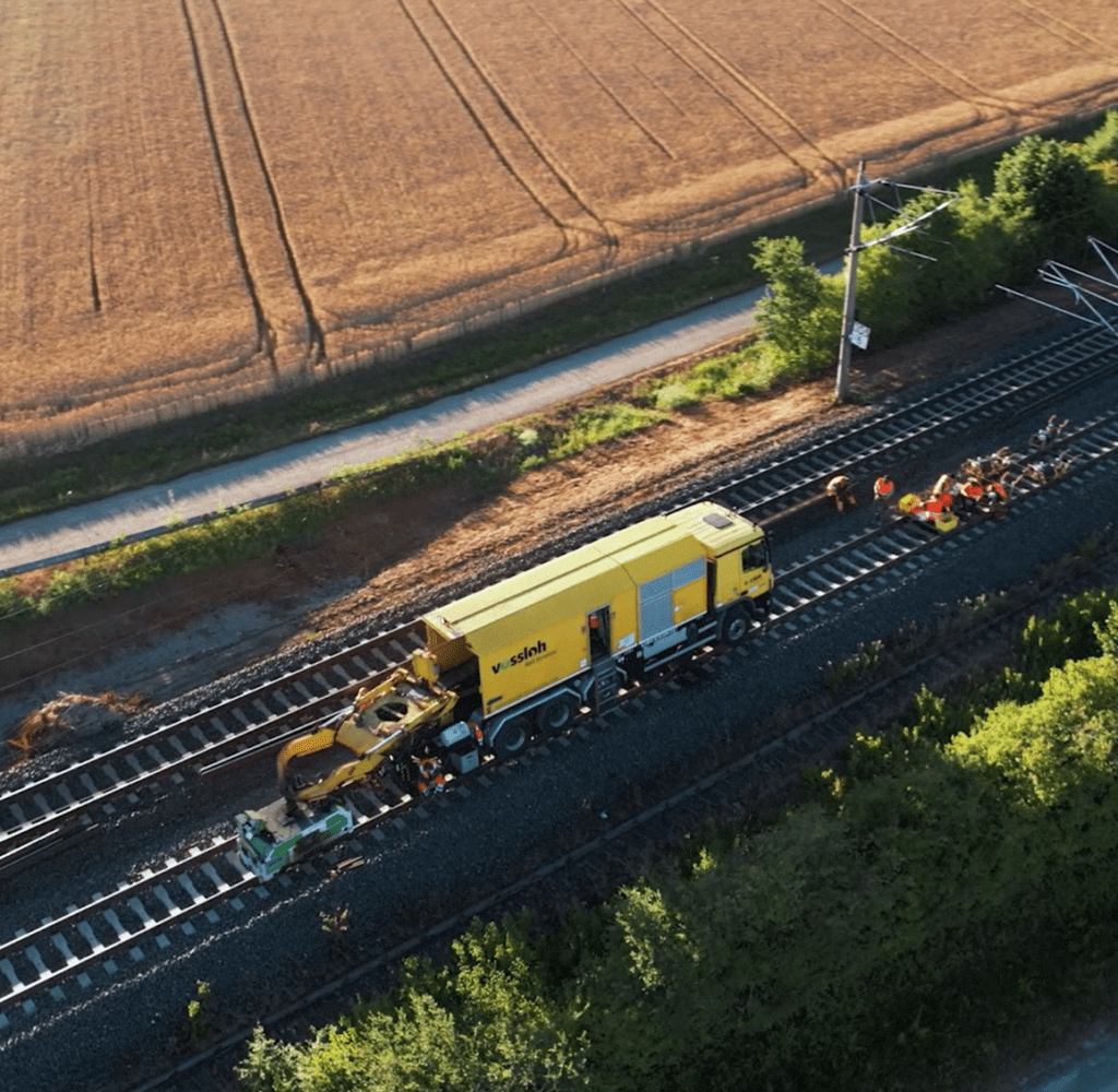 Aerial view of a yellow rail maintenance machine traveling along a railroad track through rural farmland, representing infrastructure projects and industrial supply chains.