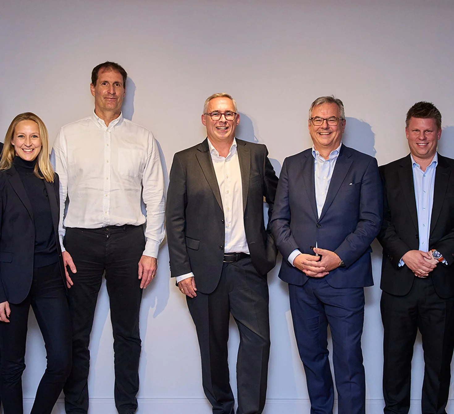 Group portrait of five business professionals dressed in suits standing against a plain wall, representing leadership team collaboration in consulting